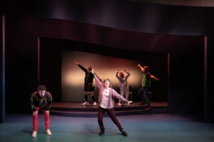 A young man in a colorful sweater leans over clasping his knees. His grandparents are wearing headbands and doing a Jazzercise routine next to him.