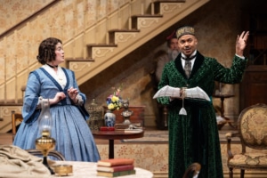 A woman with curly brown hair, in a blue, 19th century style skirt looks at a black man in a green patterned robe, fez, and cravat who is holding a model airplane.