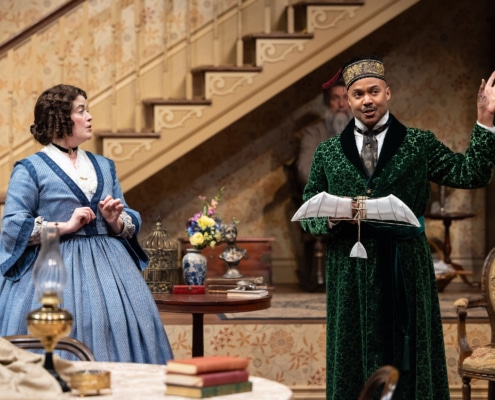 A woman with curly brown hair, in a blue, 19th century style skirt looks at a black man in a green patterned robe, fez, and cravat who is holding a model airplane.