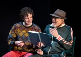 A young man in a colorful sweater sits on a park bench next to his grandfather who is wearing a green track suit and a fedora. They are laughing and reading a book together