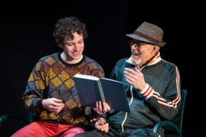 A young man in a colorful sweater sits on a park bench next to his grandfather who is wearing a green track suit and a fedora. They are laughing and reading a book together