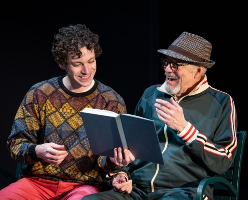 A young man in a colorful sweater sits on a park bench next to his grandfather who is wearing a green track suit and a fedora. They are laughing and reading a book together