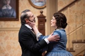 A woman with curly brown hair, in a blue, 19th century style skirt caresses the face of a man in a black 19th century suit as he holds her arm.