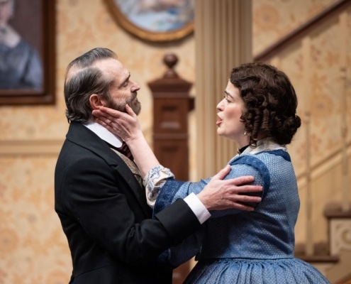 A woman with curly brown hair, in a blue, 19th century style skirt caresses the face of a man in a black 19th century suit as he holds her arm.