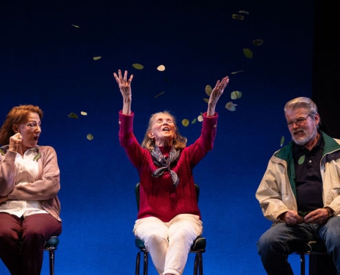 An older woman in a purple cardigan looks shocked as the older woman wearing a read sweater who is sitting next to her tries to catch spinach leaves that are falling around them like confetti. An older man in a tan windbreaker sits next to them chuckling.