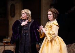 A young girl with curly blonde hair, wearing an intricate purple velvet robe and an older woman, her mother, in a bright yellow frock look concerned.