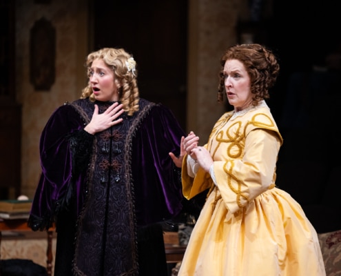 A young girl with curly blonde hair, wearing an intricate purple velvet robe and an older woman, her mother, in a bright yellow frock look concerned.