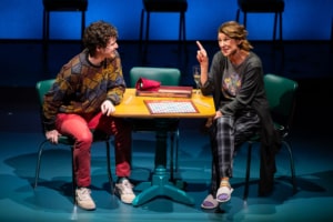 A young man in a colorful sweater grins at the older woman in pajama pants, his mother, sitting across from him. There is a game of Scrabble sitting on the table between them.