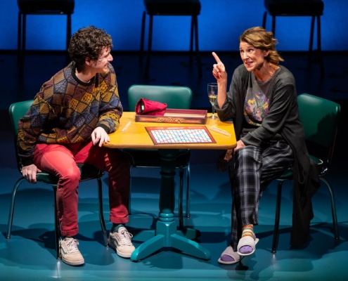 A young man in a colorful sweater grins at the older woman in pajama pants, his mother, sitting across from him. There is a game of Scrabble sitting on the table between them.