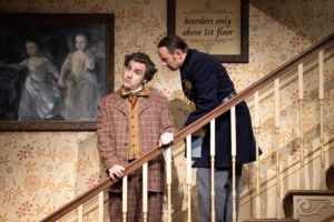 A young man in a brown plaid suit and yellow waist coat looks confused as a man in an old-fashioned police uniform speaks to him. They are standing on a staircase the constable a step above the young man.