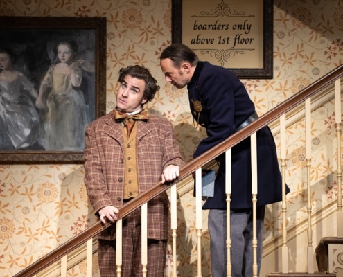 A young man in a brown plaid suit and yellow waist coat looks confused as a man in an old-fashioned police uniform speaks to him. They are standing on a staircase the constable a step above the young man.