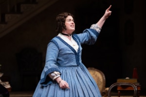 A woman with curly brown hair, in a blue, 19th century style skirt points up in the air looking determined.