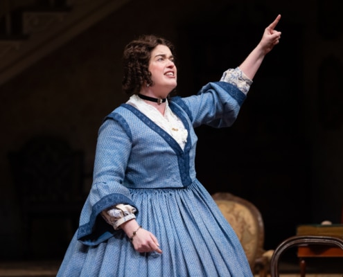 A woman with curly brown hair, in a blue, 19th century style skirt points up in the air looking determined.
