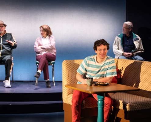 A young man in a blue and white stripped t shirt sits at a booth, he is smiling at the audience. Behing him three of his grandparents, his grandfathers and one of his grandmothers sit.