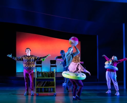 A young man in a colorful sweater stands next to a book cart his arms spread out wide. His four grandparents frolic around him, they are all wearing or playing with pool floaties.