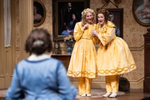 A woman with curly brown hair, in a blue, 19th century style skirt looks at a young blonde woman and her mother as they smile at her and clutch one another excitedly. They are both wearing bright yellow frocks and bloomers.