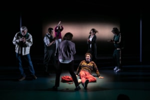 A young man in a colorful sweater lays on the ground, he is illuminated with a spotlight, and he is smiling. Six figures shrouded in darkness surround him and applaud.