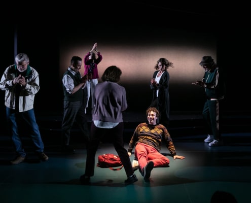 A young man in a colorful sweater lays on the ground, he is illuminated with a spotlight, and he is smiling. Six figures shrouded in darkness surround him and applaud.