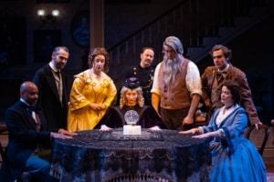 A young blonde woman in a intricate purple robe and cap looks at a glass orb on the table. A group of characters dressed in 19th century clothes surround the table and look on curiously.