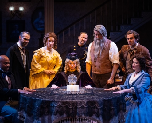 A young blonde woman in a intricate purple robe and cap looks at a glass orb on the table. A group of characters dressed in 19th century clothes surround the table and look on curiously.