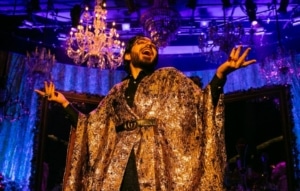Michael Shayan playing his mother Roya is dressed in an intricate gold robe and large glasses, gestures grandly.