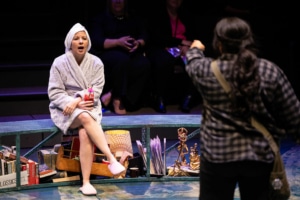 A woman in a bathrobe and slippers and a towel wrapped around her hair sits on a bench and speaks to a young woman in a black and gray flannel shirt.