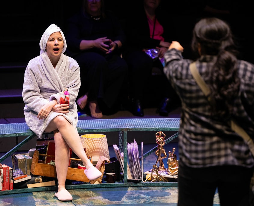 A woman in a bathrobe and slippers and a towel wrapped around her hair sits on a bench and speaks to a young woman in a black and gray flannel shirt.
