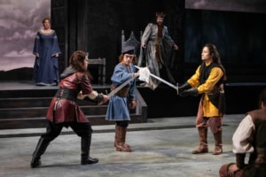 Two men with long braided hair, dressed in medieval tunics prepare for a swordfight. Their swords are crossed and third man in medieval clothes acts as referee.