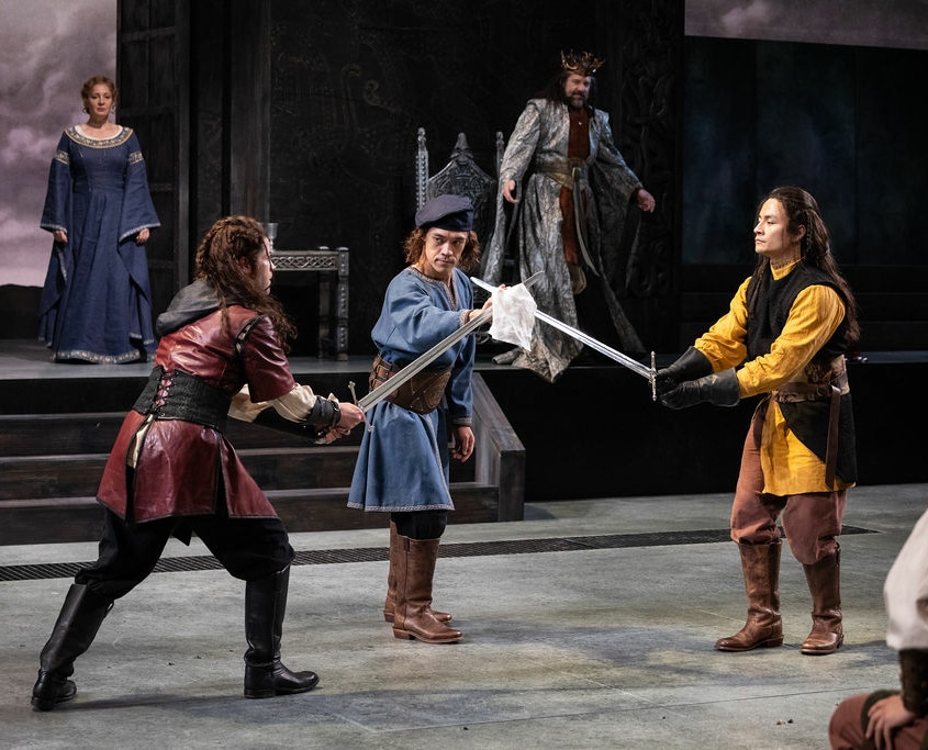 Two men with long braided hair, dressed in medieval tunics prepare for a swordfight. Their swords are crossed and third man in medieval clothes acts as referee.