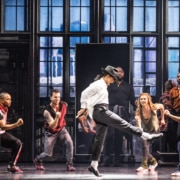 Michael Jackson dances in rehearsal, wearing the signature black and white ensemble. Backup dancers join in, facing him.