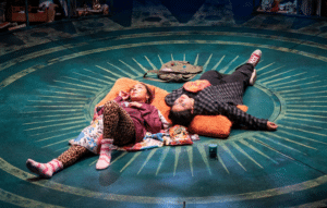 Two teenage girls lounge against a fluffy orange body pillow, eating Flamin’ Hot Cheetos. One is wearing fuzzy socks, cheetah leggings, and a red hoodie. The other is wearing red converse, black jeans, a band t-shirt, and a black and gray flannel.
