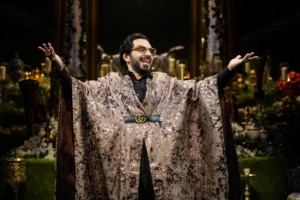 Michael Shayan playing his mother Roya is dressed in an intricate gold robe and large glasses, gestures grandly.