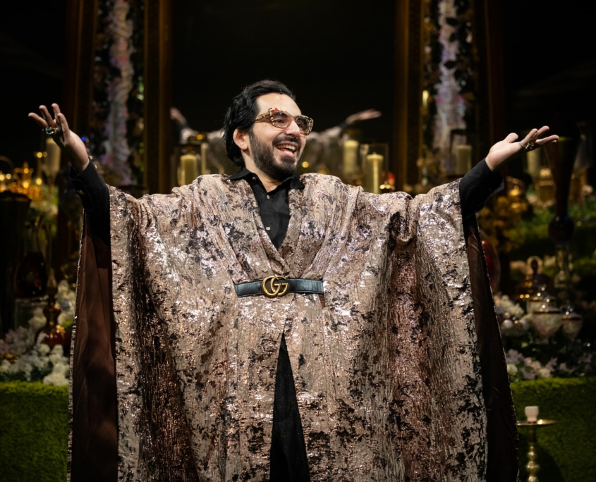 Michael Shayan playing his mother Roya is dressed in an intricate gold robe and large glasses, gestures grandly.