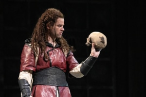 A young man with long braided hair is dressed in a red tunic with black leather arm guards. Her holds a human skull in his hand so that it faces him.