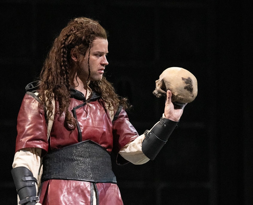 A young man with long braided hair is dressed in a red tunic with black leather arm guards. Her holds a human skull in his hand so that it faces him.
