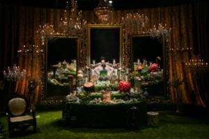 Michael Shayan playing his mother Roya is dressed in an intricate gold robe and large glasses, gestures grandly.