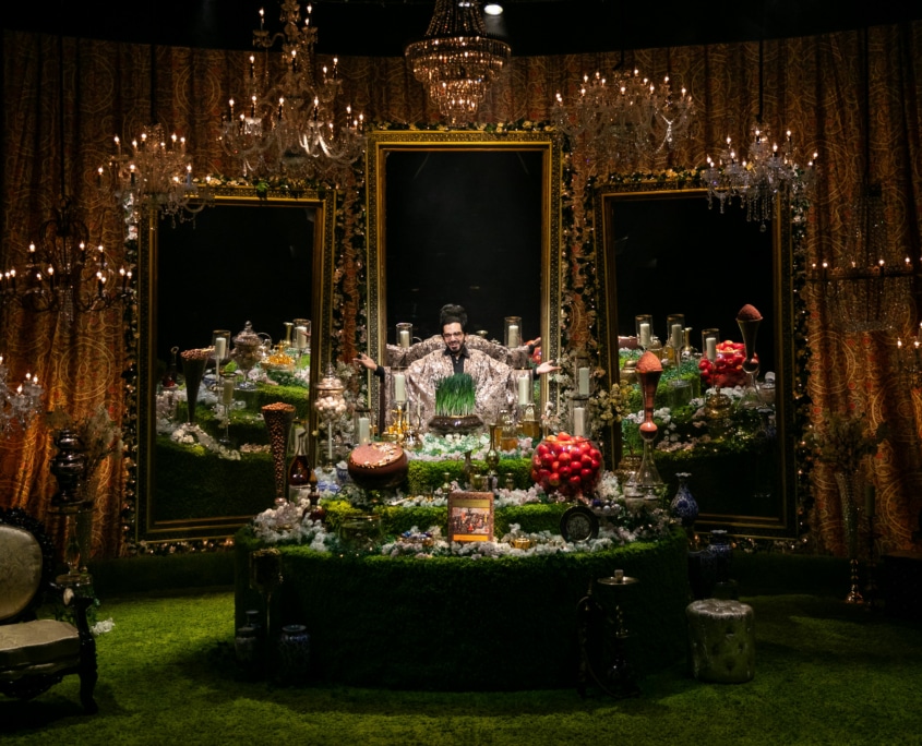 Michael Shayan playing his mother Roya is dressed in an intricate gold robe and large glasses, gestures grandly.