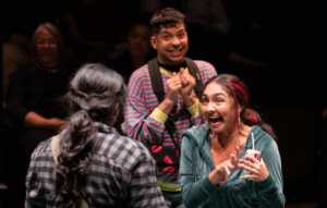 A teenage girl wearing a black and gray flannel faces a two other teenagers in colorful clothing who are gesturing excitedly.