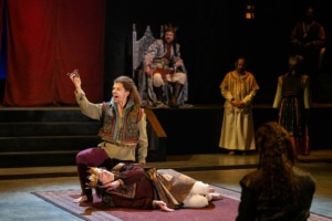 A man in a king costume with a gold crown lays on his side miming sleep. A man kneels above him in colorful clothes holding a small bottle of poison. A group of people in medieval dress watch the play they are performing.