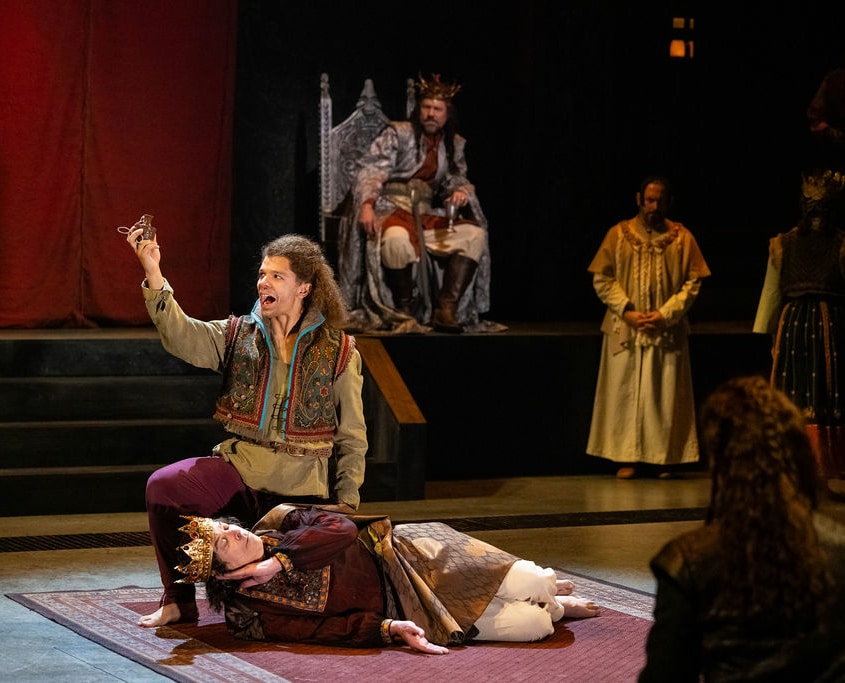 A man in a king costume with a gold crown lays on his side miming sleep. A man kneels above him in colorful clothes holding a small bottle of poison. A group of people in medieval dress watch the play they are performing.
