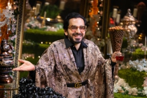 Michael Shayan playing his mother Roya is dressed in an intricate gold robe and large glasses, gestures grandly.