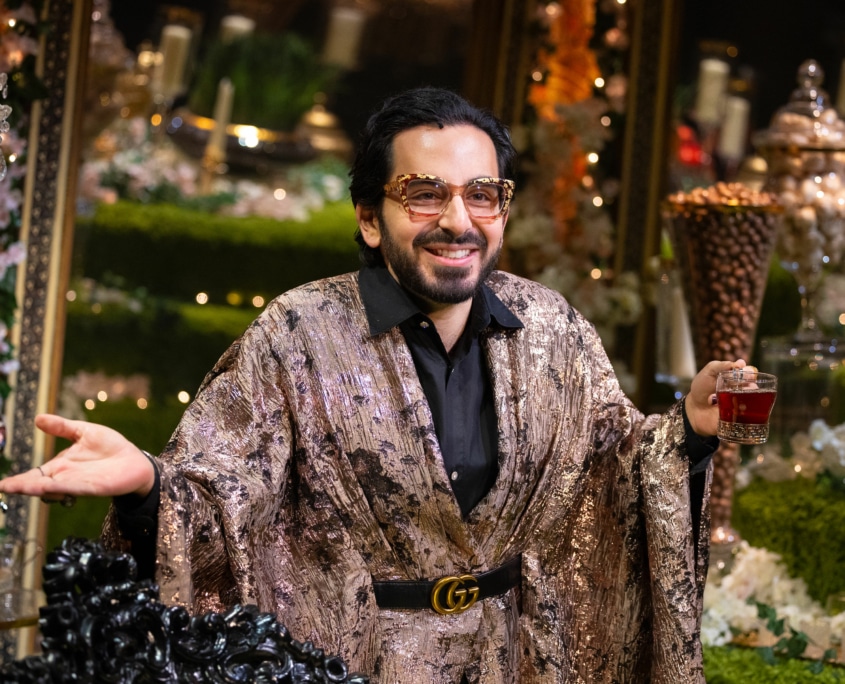 Michael Shayan playing his mother Roya is dressed in an intricate gold robe and large glasses, gestures grandly.