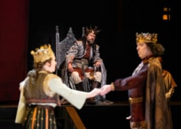 A man in a silver cloak wearing a crown of gold branches, sits on his throne looking concerned as he watches a man and woman wearing golden crowns perform in the foreground.