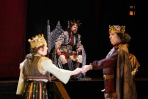 A man in a silver cloak wearing a crown of gold branches, sits on his throne looking concerned as he watches a man and woman wearing golden crowns perform in the foreground.