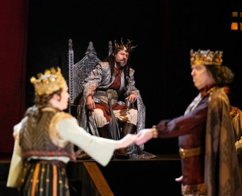 A man in a silver cloak wearing a crown of gold branches, sits on his throne looking concerned as he watches a man and woman wearing golden crowns perform in the foreground.
