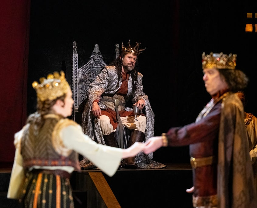 A man in a silver cloak wearing a crown of gold branches, sits on his throne looking concerned as he watches a man and woman wearing golden crowns perform in the foreground.