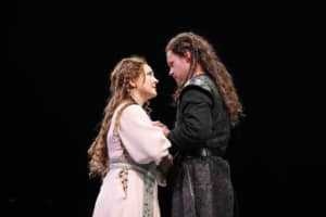 A woman with long braided hair wearing a pink dress and a man with long braided hair and a black tunic grasp one another’s hands and look intently at each other.
