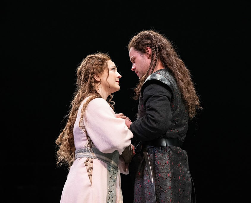 A woman with long braided hair wearing a pink dress and a man with long braided hair and a black tunic grasp one another’s hands and look intently at each other.