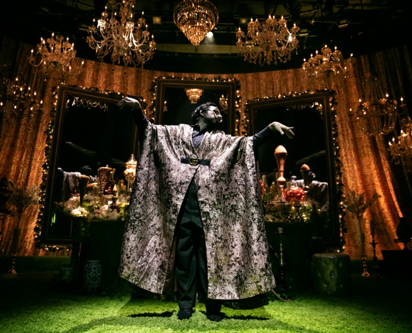 Michael Shayan playing his mother Roya is dressed in an intricate gold robe and large glasses, gestures grandly.