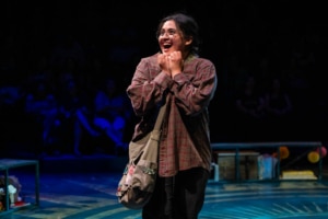 A teenage girl in a red and tan flannel and wearing round glasses grins excitedly